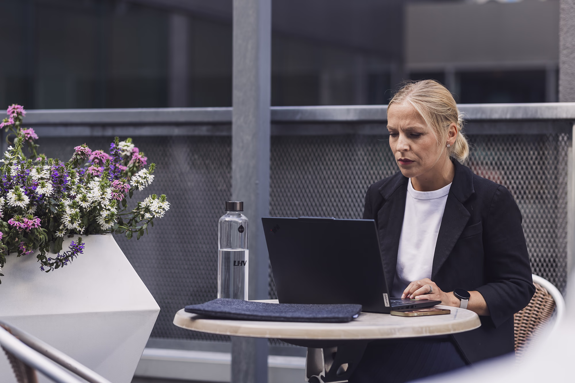 A professional works on a laptop at an outdoor table beside flowers, a water bottle, and a laptop sleeve.