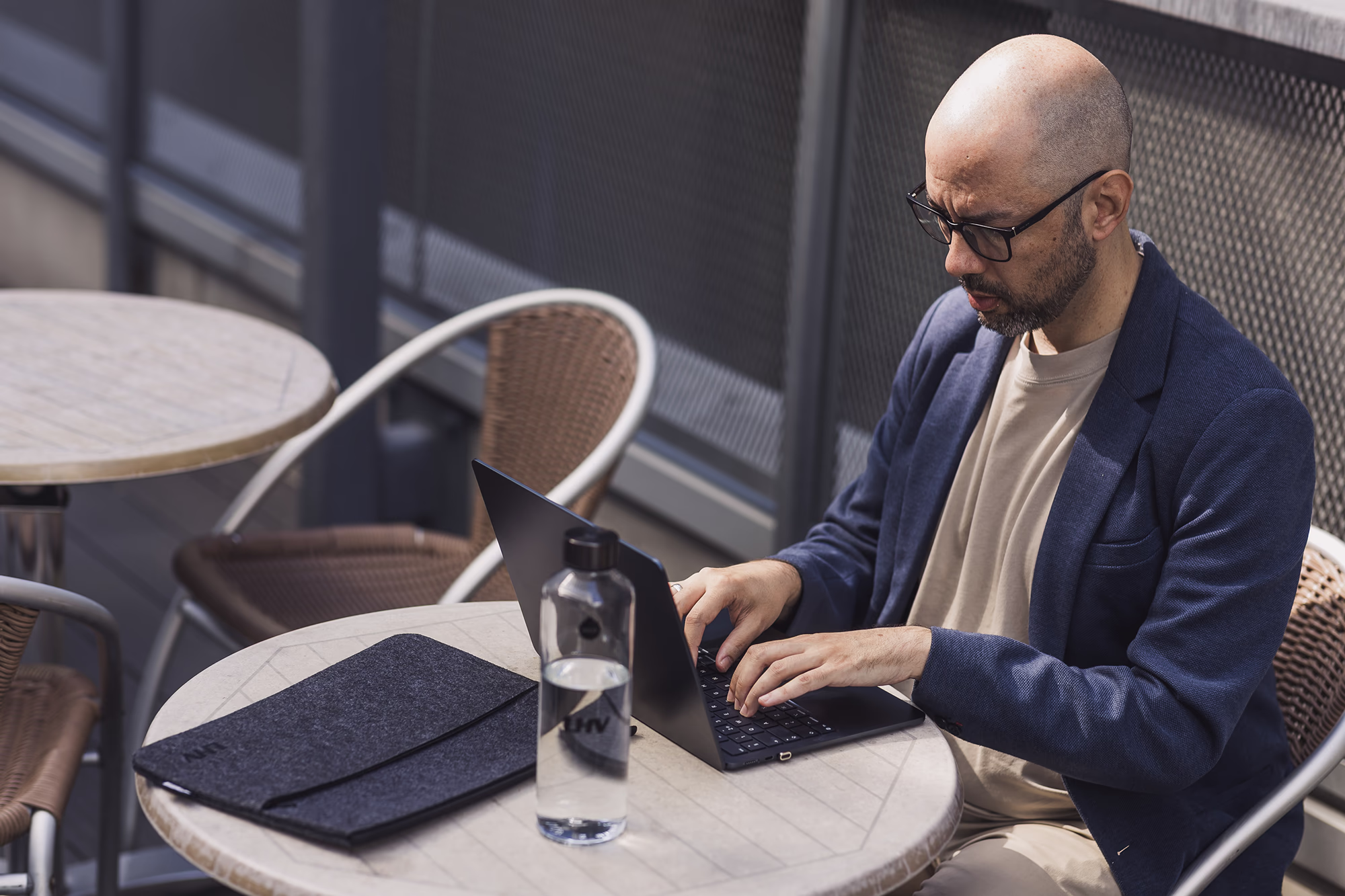 A focused man types on a laptop at an outdoor table with a water bottle and laptop sleeve nearby.